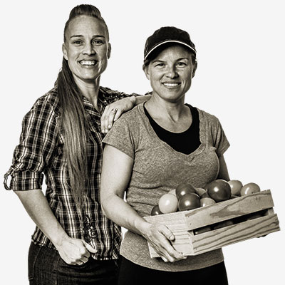 two women standing next to each other while one holds a crate of tomatoes